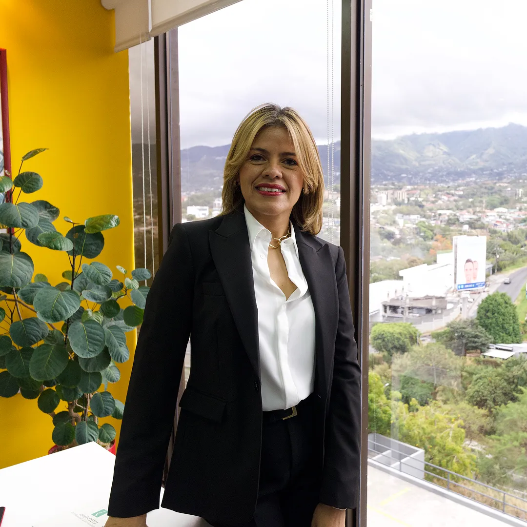 Una mujer ejecutiva sonriente con traje sastre negro y camisa blanca, de pie en una oficina corporativa junto a un gran ventanal con vistas a una ciudad y montañas de fondo.
